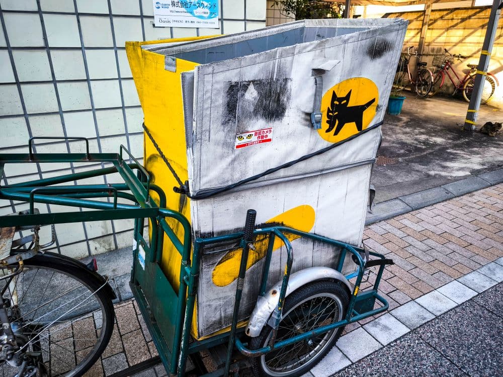 Ein verwitterter Lieferwagen, der an einem Fahrrad mit dem schwarzen Katzenlogo des Takkyubin-Dienstes von Yamato Transport befestigt ist, steht auf einem gefliesten Gehweg neben einem Gebäude und mehreren anderen Fahrrädern.