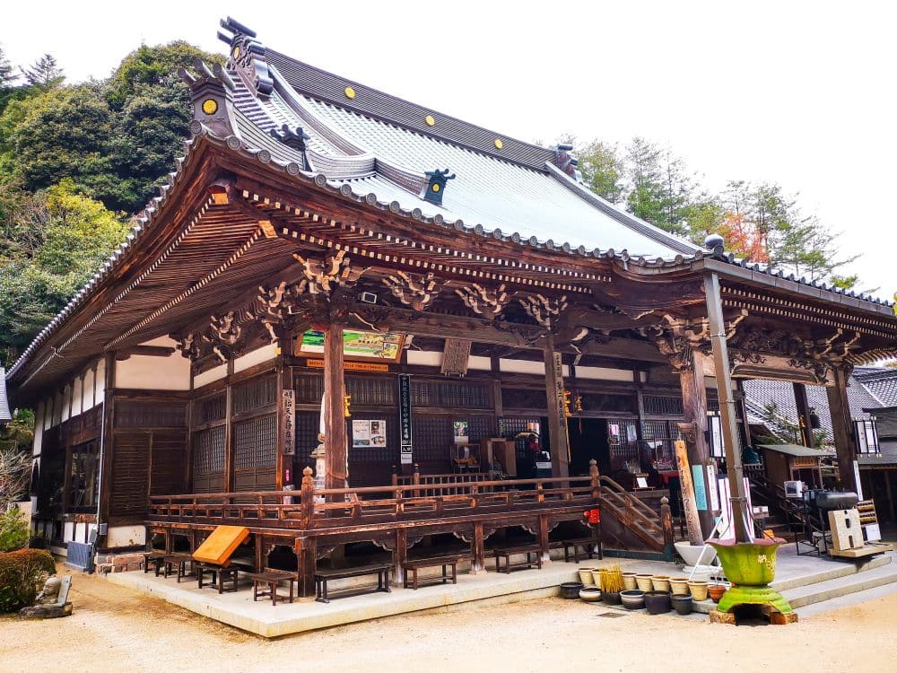 Ein traditioneller japanischer Tempel, Daishoin, mit kunstvollen Holzschnitzereien und einem Ziegeldach, umgeben von Bäumen. In der Nähe des Eingangs sind Menschen zu sehen, im Vordergrund stehen Topfpflanzen und Bänke.