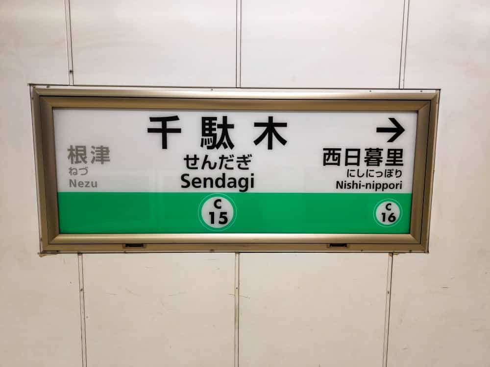 Ein Schild der japanischen U-Bahn zeigt die Station Sendagi (C15) in der Mitte, mit Nezu (C14) auf der linken Seite und Nishi-nippori (C16) auf der rechten Seite. Die Stationsnamen werden sowohl auf Japanisch als auch auf Englisch angezeigt.