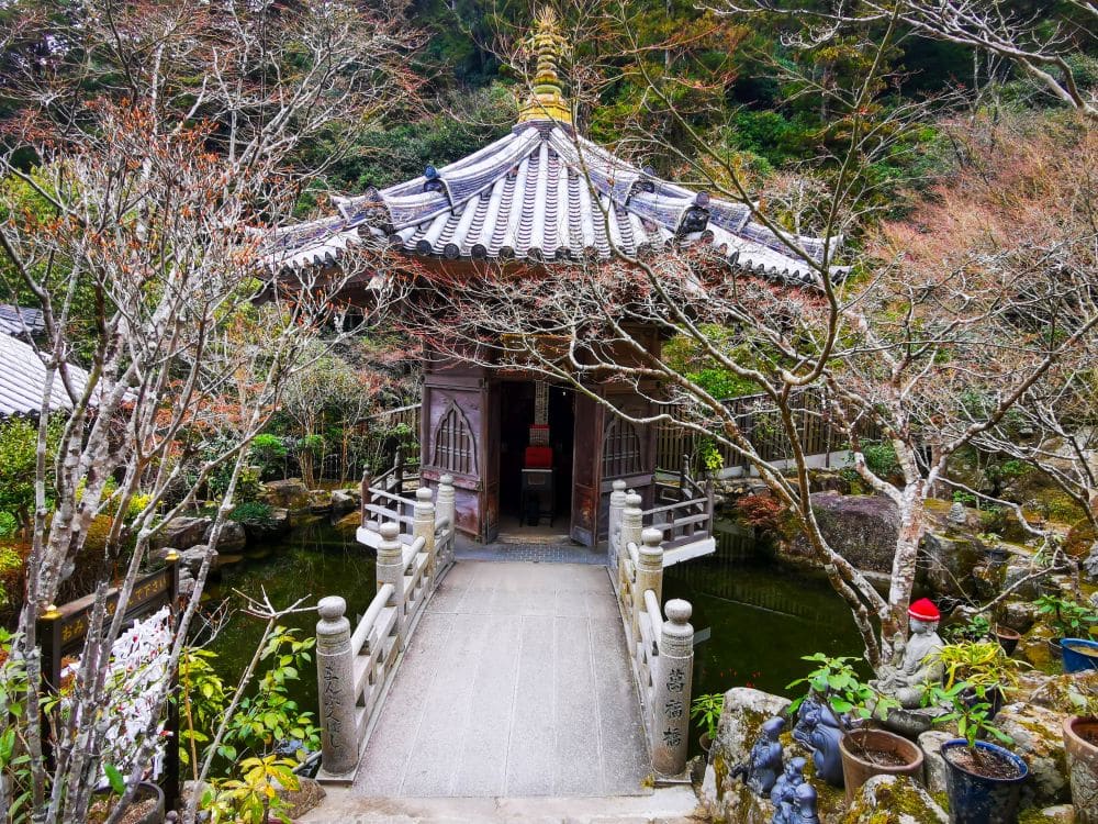 Ein traditioneller japanischer Pavillon mit Ziegeldach steht über einem Teich, zu dem man über eine kleine Steinbogenbrücke gelangt, umgeben von kahlen Bäumen und üppigem Grün in der ruhigen Gartenanlage Daishoin.
