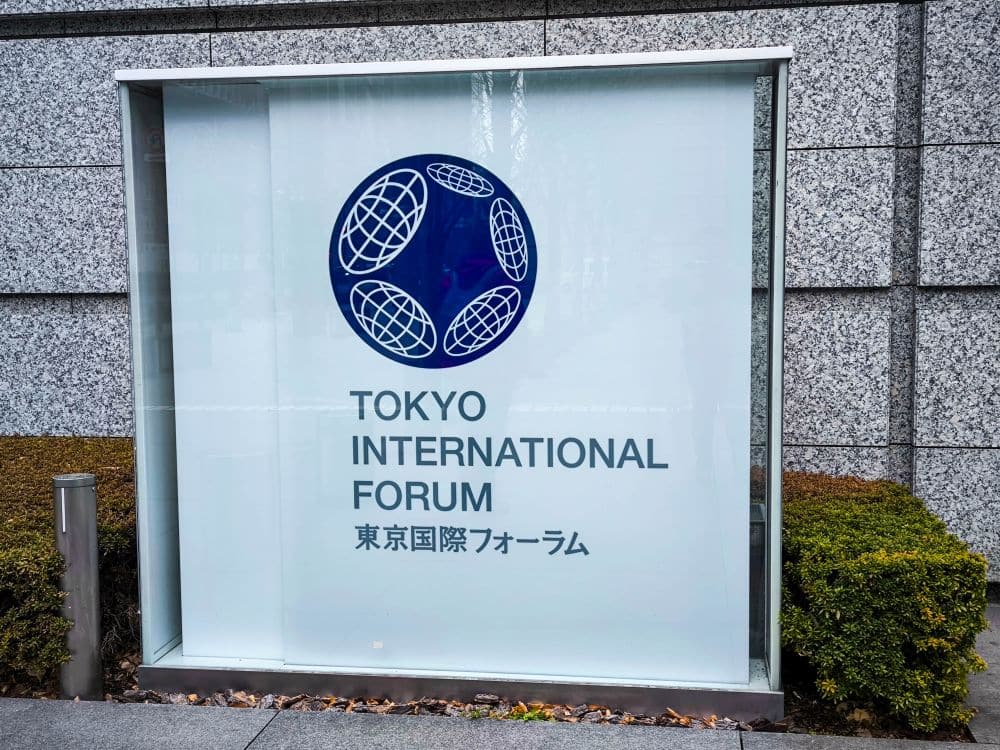 Ein Schild des Tokyo International Forum zeigt das blaue Logo über englischem und japanischem Text auf einer Glasscheibe vor dem eleganten, modernen Gebäude, das von grauen Steinmauern und üppigen grünen Sträuchern eingerahmt wird.