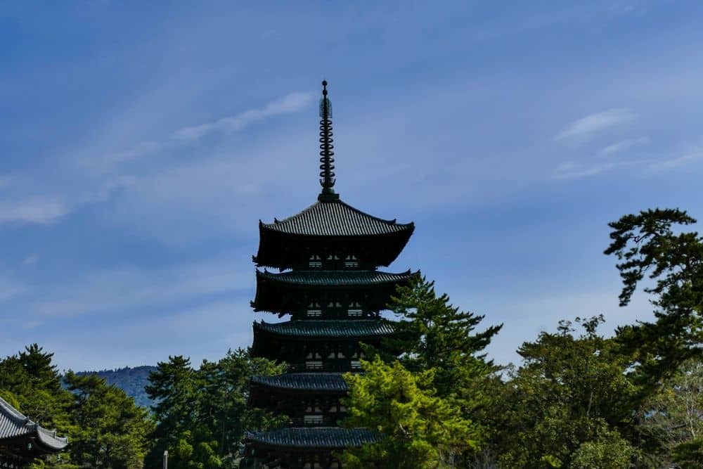 Die traditionelle fünfstöckige Pagode des Kōfuku-ji-Tempels erhebt sich inmitten grüner Bäume in Nara unter einem blauen Himmel. Ihre dunkle Silhouette erinnert an eine Zeitreise und steht in schönem Kontrast zur hellen Naturumgebung.