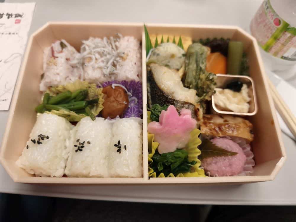 Eine japanische Bento-Box mit Reisbällchen, eingelegter Pflaume, Fisch, Gemüse, Tempura und einer rosafarbenen Blume, die ordentlich in Fächern angeordnet ist - ein perfektes Konbini-Frühstück für Japanreisende. Im Hintergrund sind ein Getränk und Essstäbchen zu sehen.