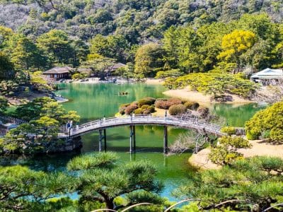 Ein malerischer japanischer Garten in Takamatsu mit der Gartenkunst im Ritsurin-Garten mit einer geschwungenen Holzbrücke über einen Teich, umgeben von üppig grünen Bäumen, Sträuchern und traditionellen Gebäuden in hellem Sonnenlicht.