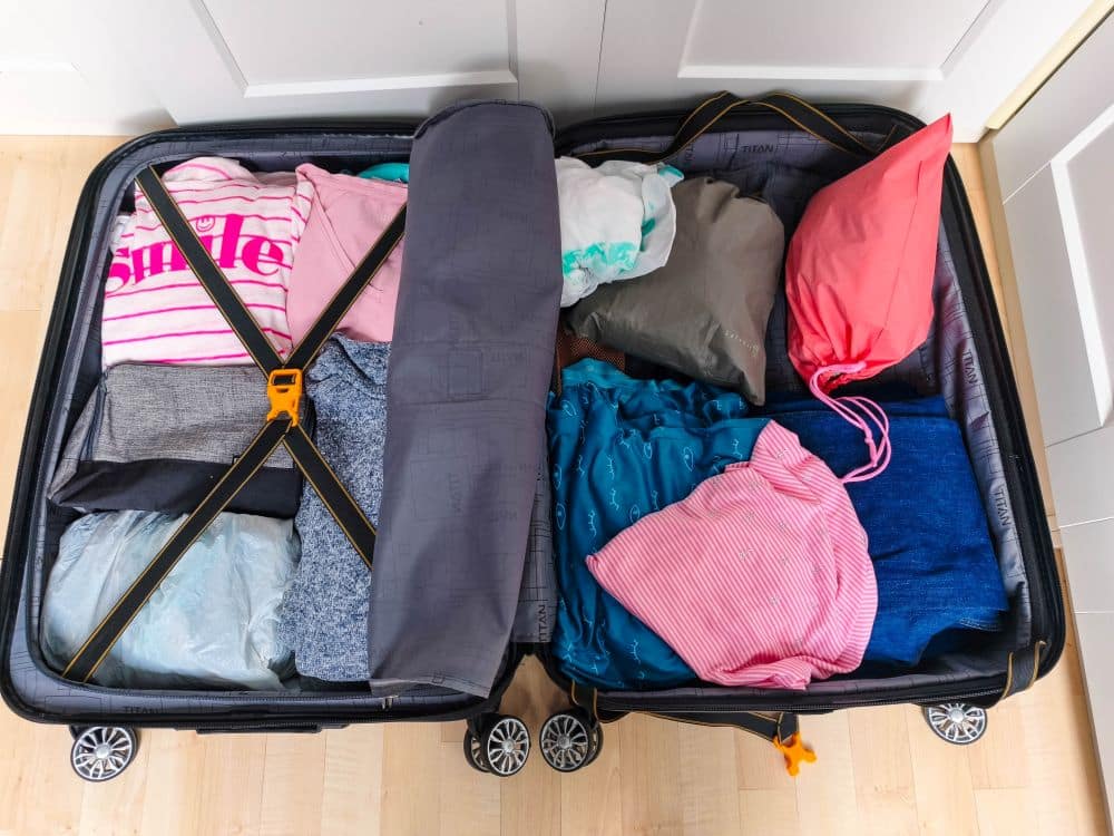 Ein offener Koffer, ordentlich gepackt mit bunten Kleidern und einem Kissen, steht auf einem hellen Holzboden vor weißen Schranktüren - das perfekte Bild für die Erstellung Ihrer nächsten Packliste.