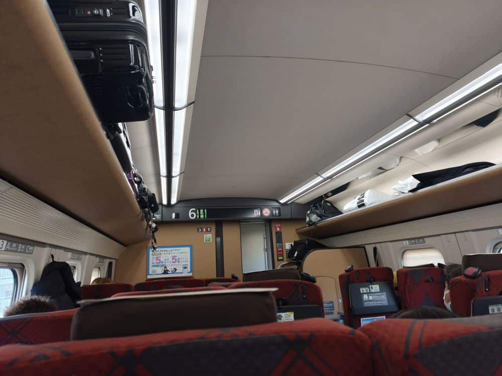 Innenansicht eines Zugwagens mit roten Sitzen, Gepäck auf Gepäckablagen und beleuchteten Schildern mit Sitzplatznummern. Die Fahrgäste entspannen sich, während ein Bildschirm die Reisedaten anzeigt - perfekt für Schienenabenteuer mit dem JR Kyushu Rail Pass.