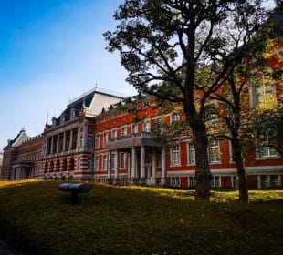 In Nagatachō, dem politischen Herzen Japans, steht an einem sonnigen Tag bei strahlend blauem Himmel ein großes rotes Backsteingebäude mit weißen Säulen und einem Mansarddach hinter gestutzten Hecken und einem Baum.
