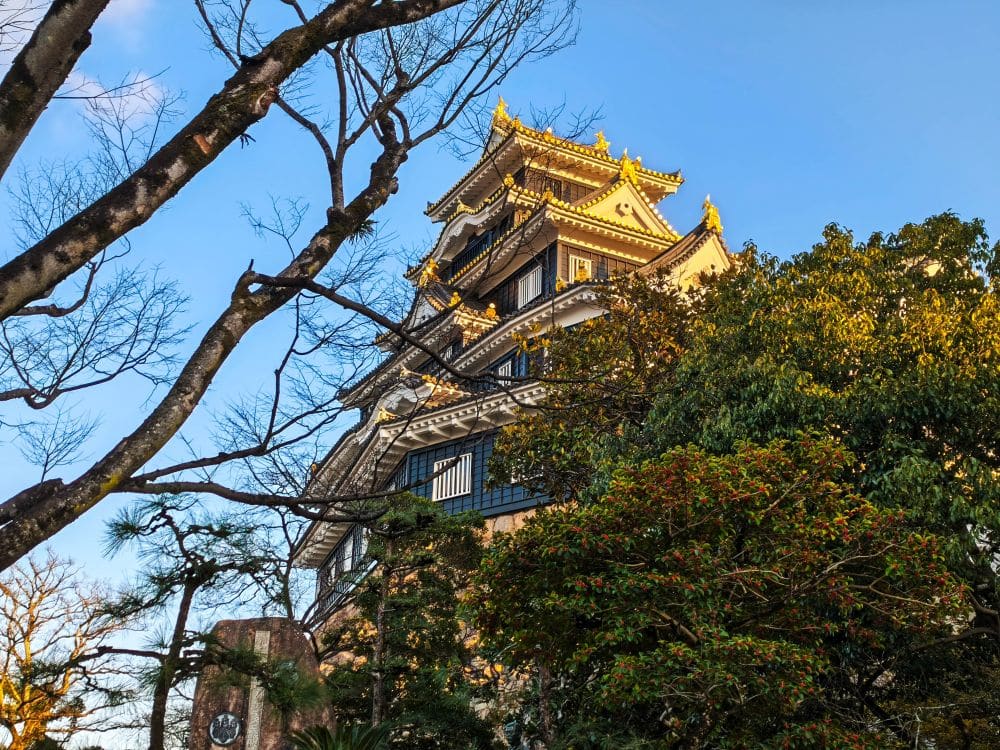 Eine traditionelle japanische Burg, bekannt als Krähenburg oder Okayama Castle, mit verzierten Dächern und goldenen Akzenten erhebt sich über grünen Bäumen, eingerahmt von kahlen Ästen unter einem klaren blauen Himmel.