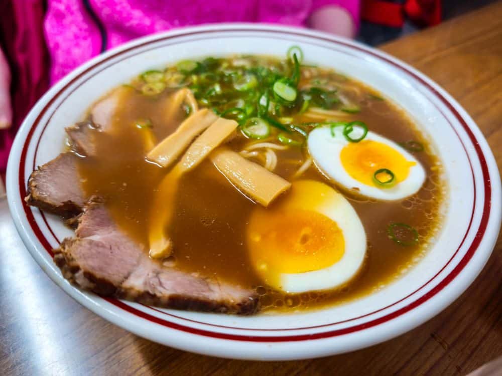 Eine Schüssel Okayama-Ramen mit Schweinefleischscheiben, Bambussprossen, Frühlingszwiebeln, Nudeln und einem halbierten weichgekochten Ei in Brühe, serviert auf einem weißen Teller mit roten Streifen, der auf einem Holztisch steht.