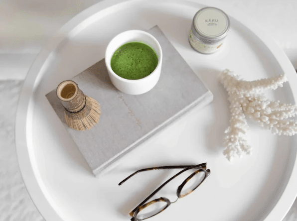 Auf einem weißen Tablett stehen eine Schale mit grünem Matcha, ein Bambusbesen, ein graues Buch, Gläser aus Schildpatt, ein Cremetopf und eine weiße Koralle - alles fein säuberlich arrangiert, um ein beruhigendes, von Matcha inspiriertes Bild zu schaffen.