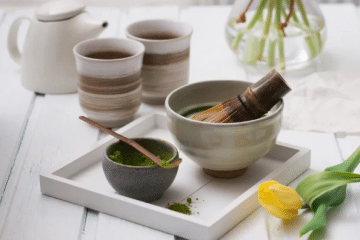 Ein Tablett mit einer Schale Japan Matcha und einem Bambusbesen, eine kleine Schale Matcha-Pulver mit einem Löffel, zwei Tassen Grüner Tee, eine weiße Teekanne, eine Vase mit Blumen und eine gelbe Tulpe auf einem weißen Holztisch.