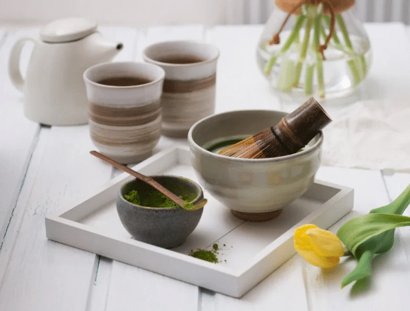Ein Tablett mit einer Schale Japan Matcha und einem Bambusbesen, eine kleine Schale Matcha-Pulver mit einem Löffel, zwei Tassen Grüner Tee, eine weiße Teekanne, eine Vase mit Blumen und eine gelbe Tulpe auf einem weißen Holztisch.
