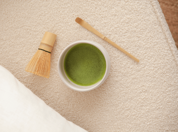 Eine Schale mit schaumigem grünem Matcha-Tee steht auf einer strukturierten Sahnefläche, links ein Bambusbesen und rechts ein Bambuslöffel, die zum beruhigenden Ritual der Matcha-Zubereitung einladen.