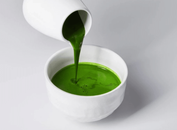 Ein weißer Krug gießt dicken, leuchtenden Matcha in eine weiße Schale, beide vor einem schlichten, hellen Hintergrund.