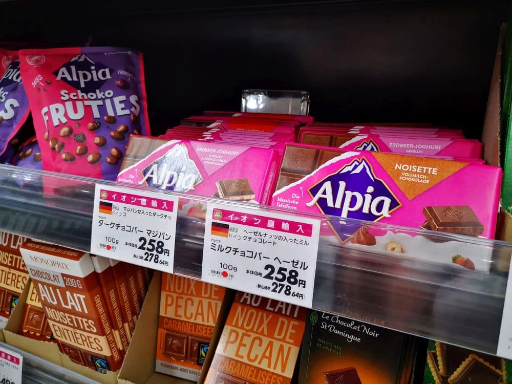 In einem Supermarktregal sind verschiedene Sorten Alpía-Schokoladentafeln und andere Pralinen mit Preisschildern in japanischen Yen ausgestellt, darunter Schoko Fruties und Nusssorten in bunten Verpackungen.