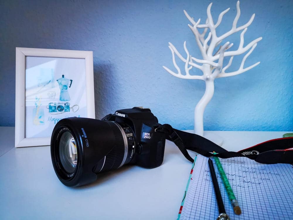 Eine Canon EOS 1200D-Kamera liegt auf einem Schreibtisch neben einem gerahmten Bild, einem weißen Dekobaum, einem Notizblock mit Gittermuster und einem grünen Bleistift - alles sehr übersichtlich und stilvoll arrangiert. Die Wand im Hintergrund ist hellblau.