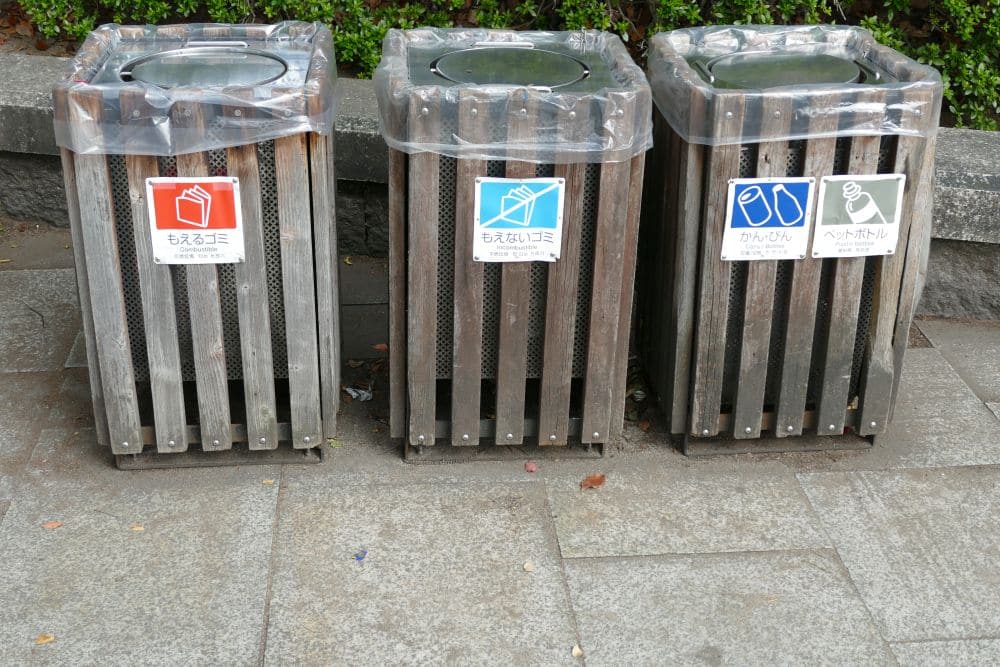 Drei hölzerne Abfalleimer mit Plastikdeckeln stehen auf einem Gehweg aufgereiht, gemäß den örtlichen Regeln für die Mülltrennung. Jede Tonne hat ein Etikett auf Japanisch für brennbare Abfälle, Dosen und Plastikflaschen. Im Hintergrund sind grüne Sträucher und eine Steinmauer zu sehen.