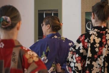 Drei Personen in bunten Kimonos stehen mit dem Rücken zur Kamera und blicken in Spiegel. Ihr Haar ist ordentlich zu einem Dutt frisiert, den jede mit einem dekorativen Accessoire befestigt hat.
