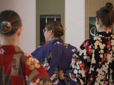 Drei Personen in bunten Kimonos stehen mit dem Rücken zur Kamera und blicken in Spiegel. Ihr Haar ist ordentlich zu einem Dutt frisiert, den jede mit einem dekorativen Accessoire befestigt hat.